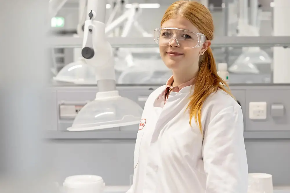 A female lab technician wearing a white, Henkel-branded coat and safety glasses smiles towards the camera. Some equipment can be seen in the background.