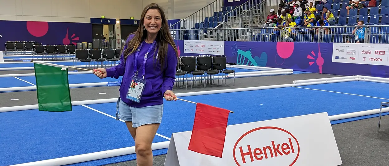 Natalia stands next to the boccia court while holding a green flag in her right hand and red flag in her left hand. 