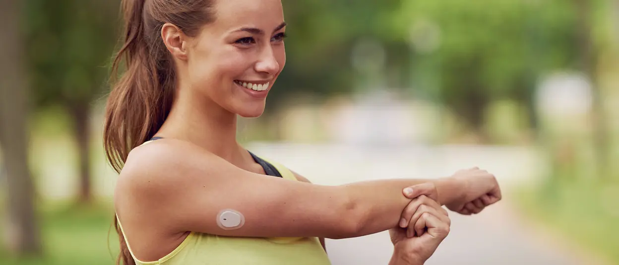 A young woman is stretching her arm preparing for exercise with a continuous glucose monitoring patch attached to her upper right arm.