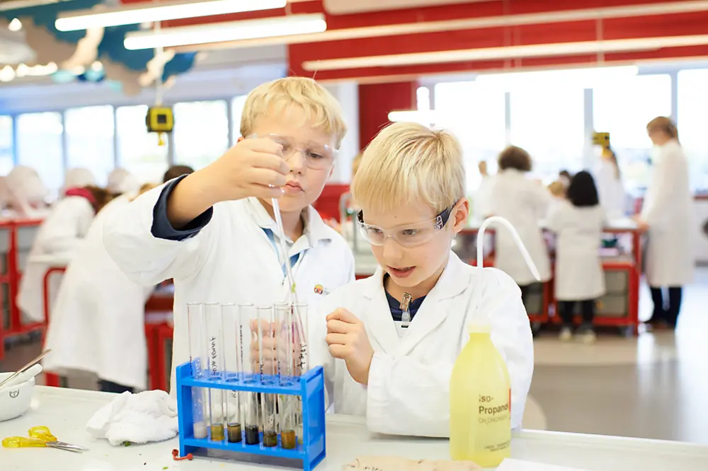 two boys in lab coats and safety glasses adding a solution to different test tubes