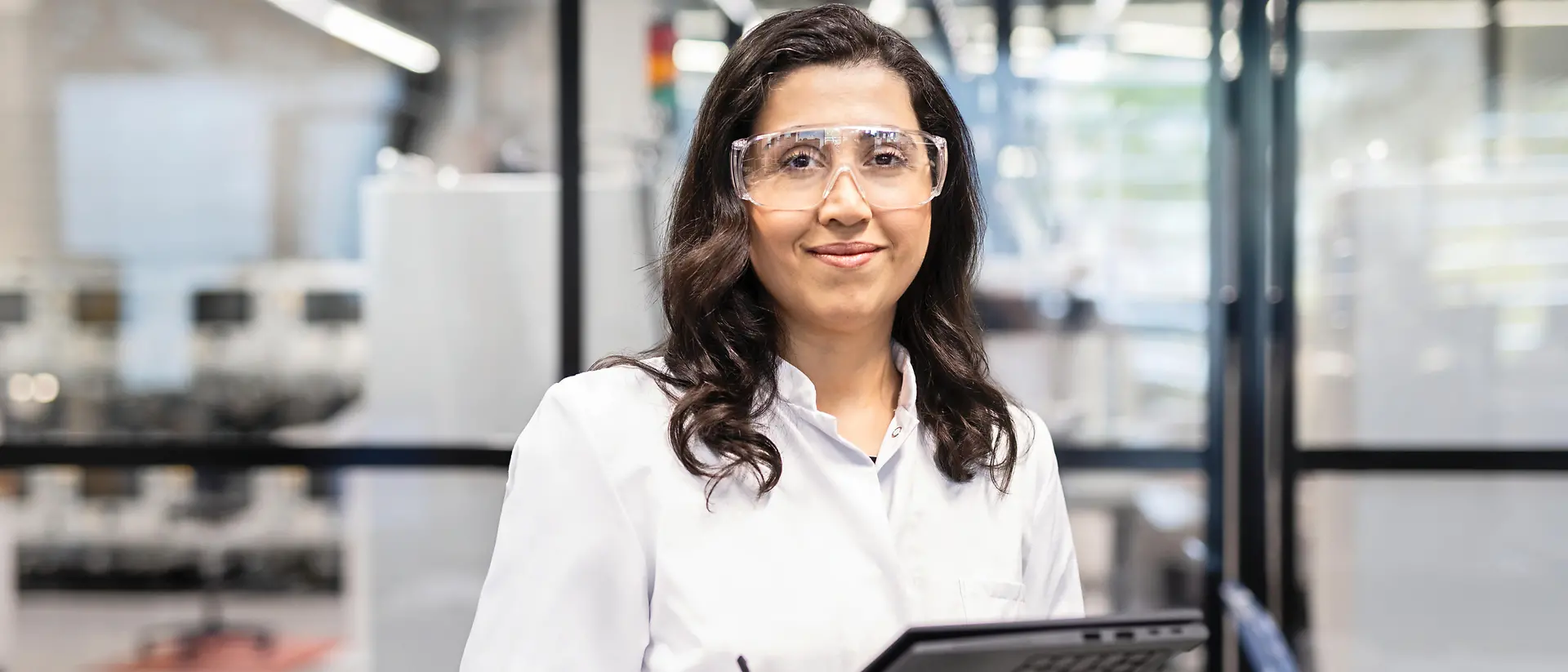 A female lab technician wearing a white coat and safety glasses and holding a tablet. She is loooking towards the camera and smiling.