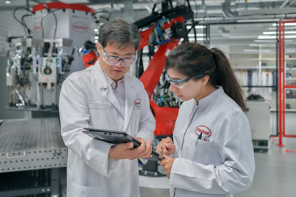 A male and a female engineers wearing white, Henkel-branded coats look at a tablet. They are inside a factory with Henkel-branded machinery.