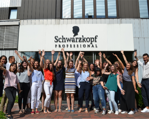 O echipă diversă Henkel care aplaudă în fața clădirii profesionale Schwarzkopf și ridică brațele