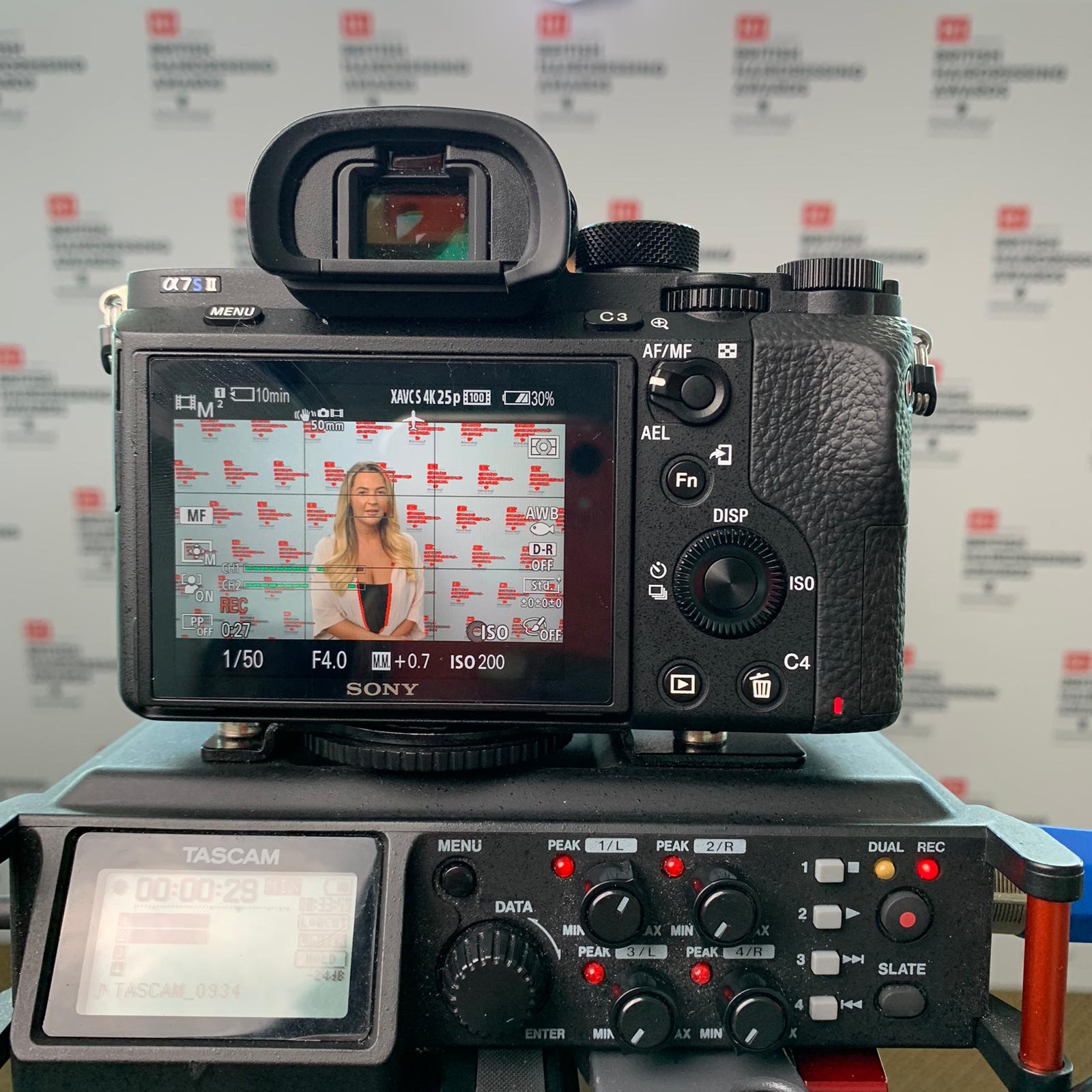 Digital camera mounted on an editing device, the display showing a woman in front of the BHA logo wall