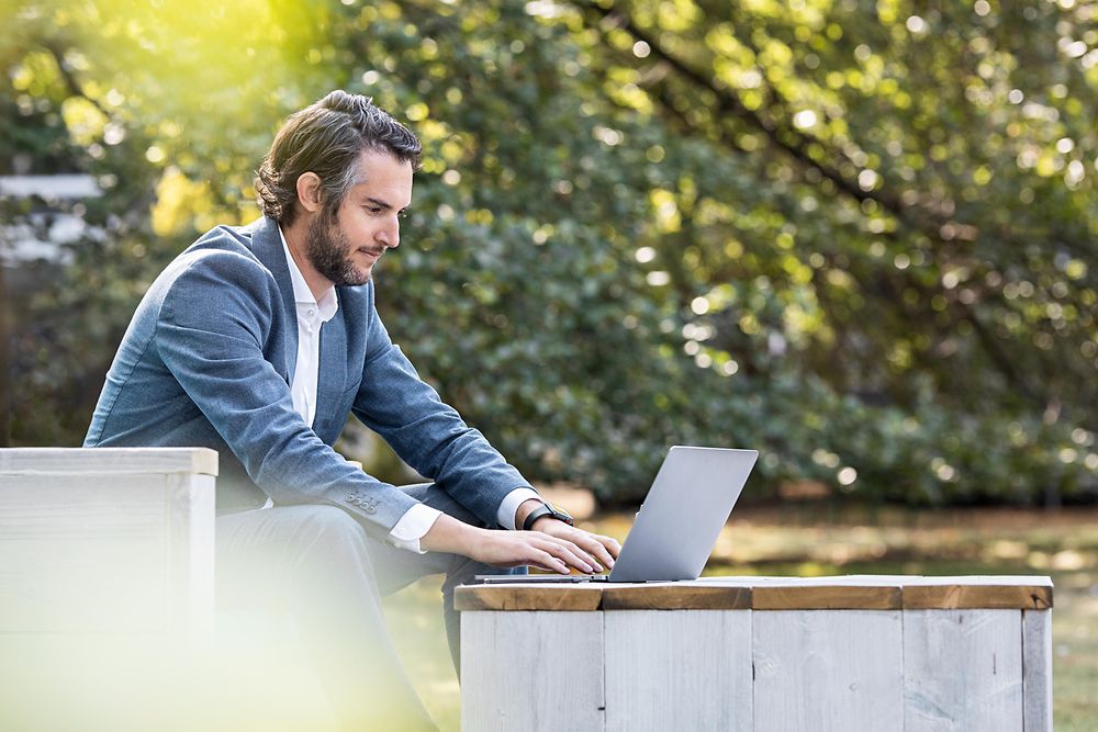 Flexible working: A man is working on his laptop outside on a sunny day, surrounded by nature.