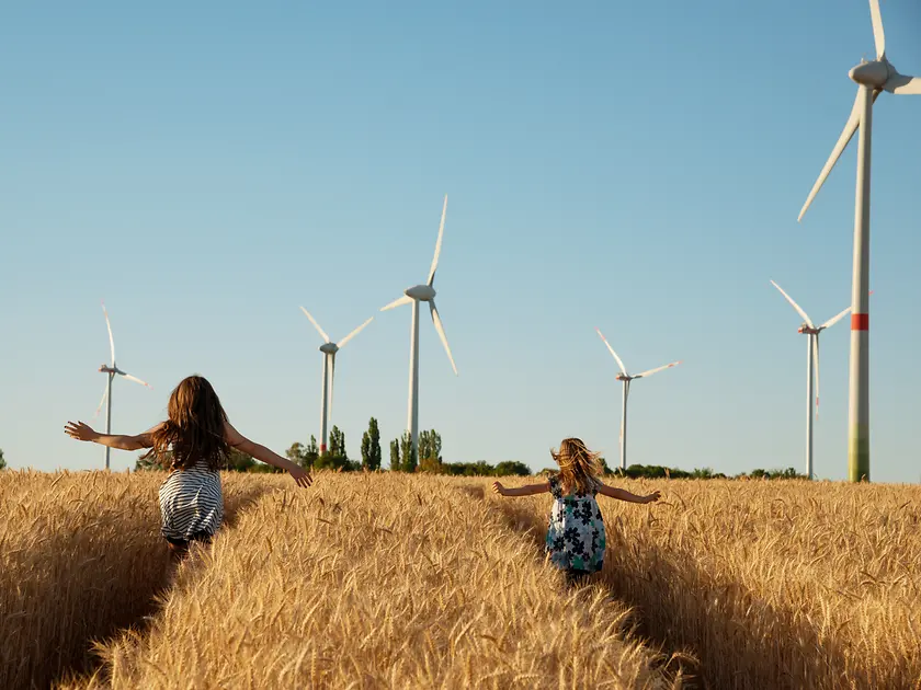 Girls run through a field towards wind turbines.