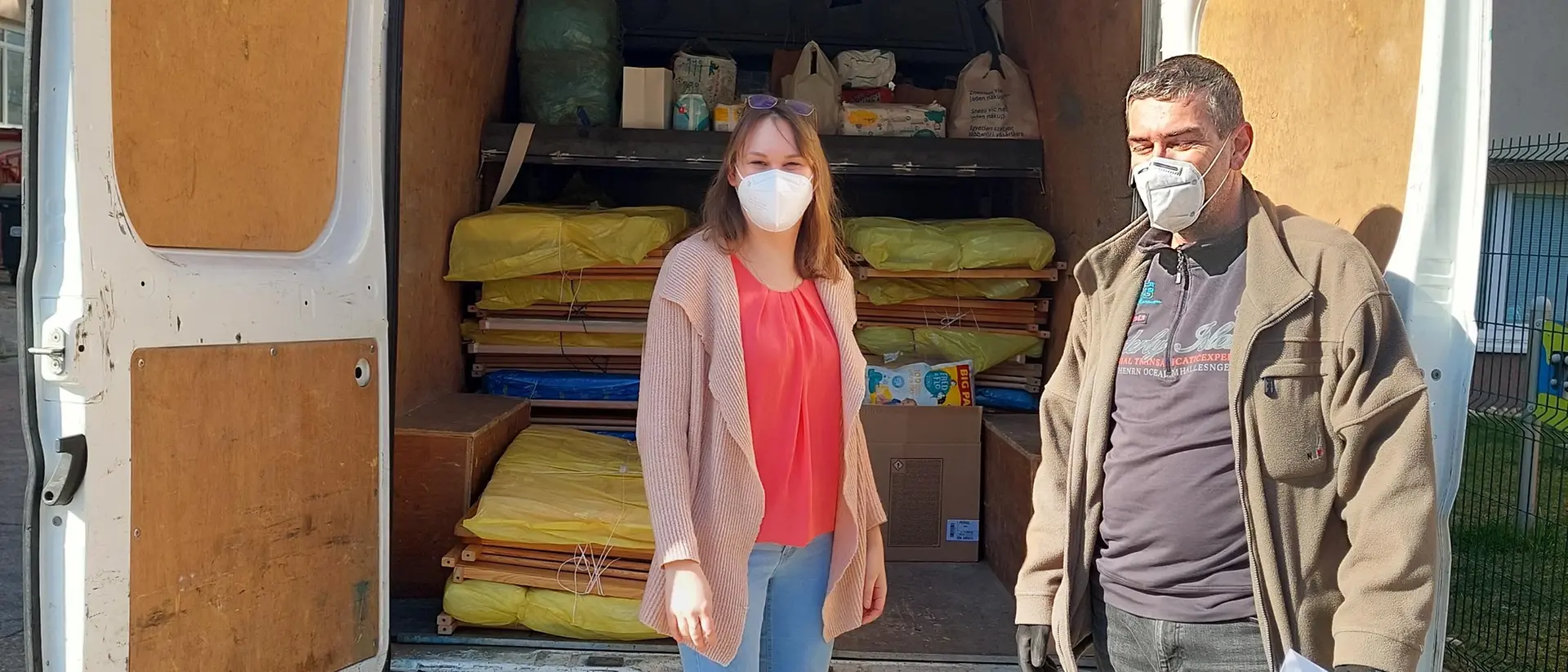 A woman and a man standing in front of a van loaded with cribs and mattresses that are being donated.