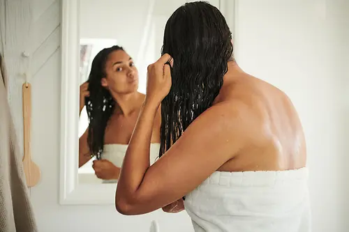 A woman stands in front of the mirror with a bath towel and wet hair.
