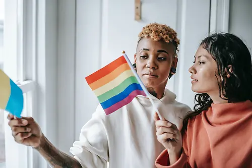 Two women are talking to each other while holding an LGBTQ+ flag.