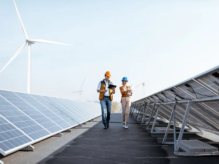 Two employees walk in between solar panels. Wind turbines are in the background. 