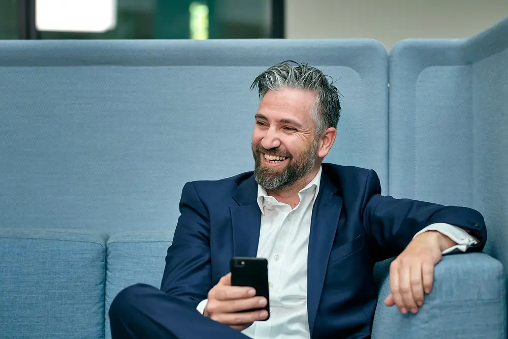 A man in a blue suit with a white shirt is sitting on a blue sofa, laughing and holding his smartphone.