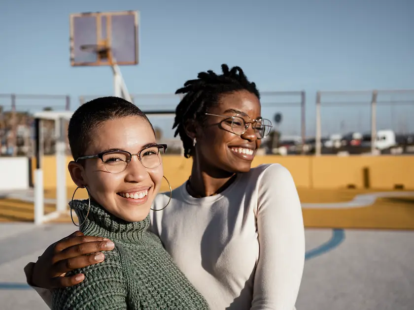 Zwei Frauen stehend lachend und umarmend auf einem Basketballplatz. 