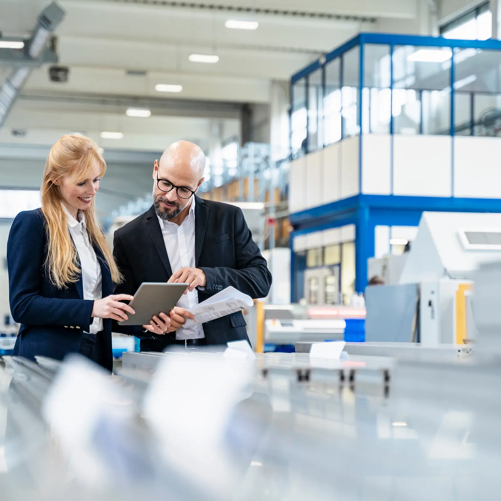 Two business people stand in a manufacturing site and look at a document