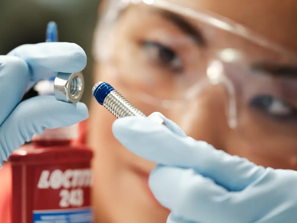 Close-up of a person with safety glassess. They have a bolt in one hand, and in the other hand a screw on which a blue LOCTITE threadlocker has been applied. The threadlocker bottle can be partially seen.