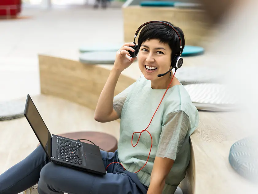 An employee has her laptop on her lap and wears a headset while smiling into the camera. 
