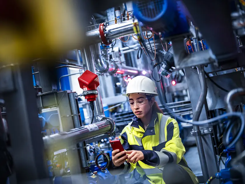 A female worker is standing in a production site checking on her mobile device the status of the equipment.
