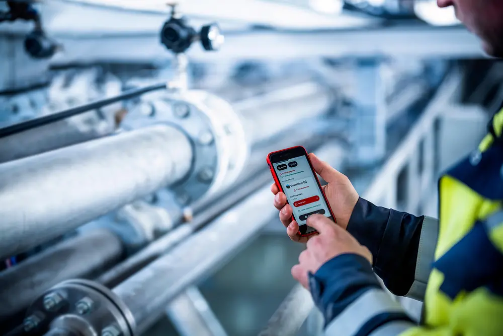 A production worker is holding a smart phone standing in front of equipment to do maintenance.