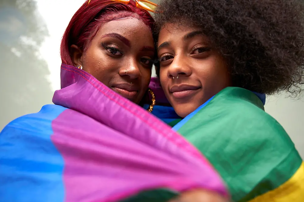 Two women look into the camera and have a pride flagged wrapped around themselves.