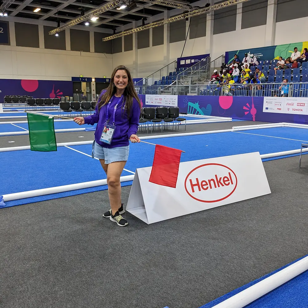 Natalia stands next to the boccia court while holding a green flag in her right hand and red flag in her left hand. 
