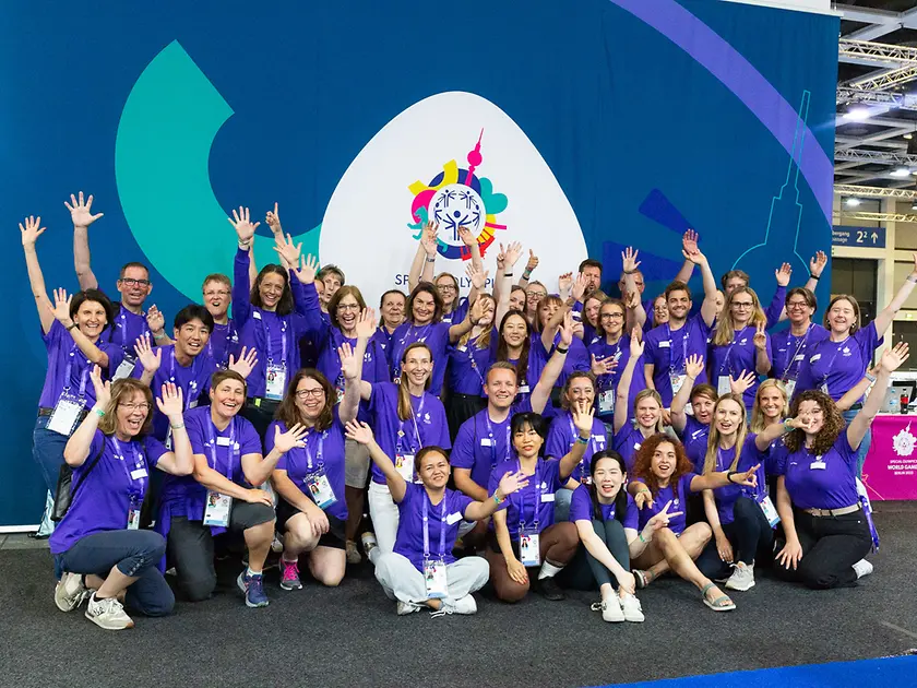 The international team of 60 Henkel volunteers wear their volunteer t-shirts and stand cheering in front of the Special Olympics World Games banner in Berlin.