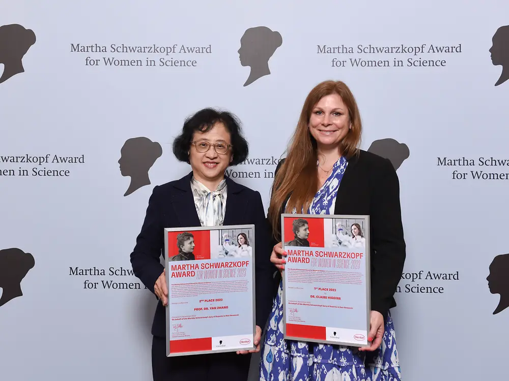 wo women standing next to each other in front of a photo wall and holding their winner's certificate. 