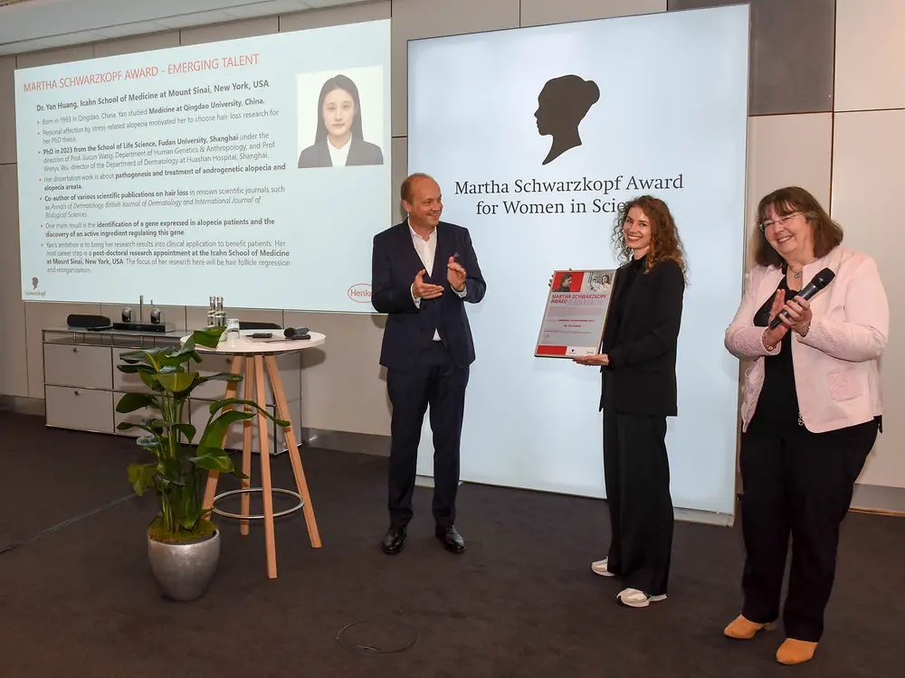 A man on the left and a woman on the right are applauding while another woman in the middle holds Dr. Yan Huang's winner's certificate. 