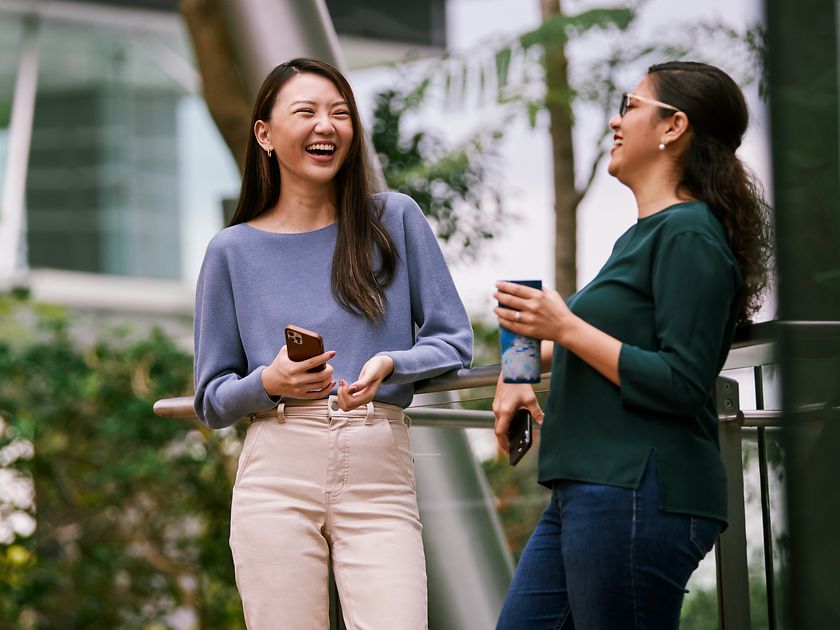 Two women standing in the green outdoor space of a modern office building laughing together.