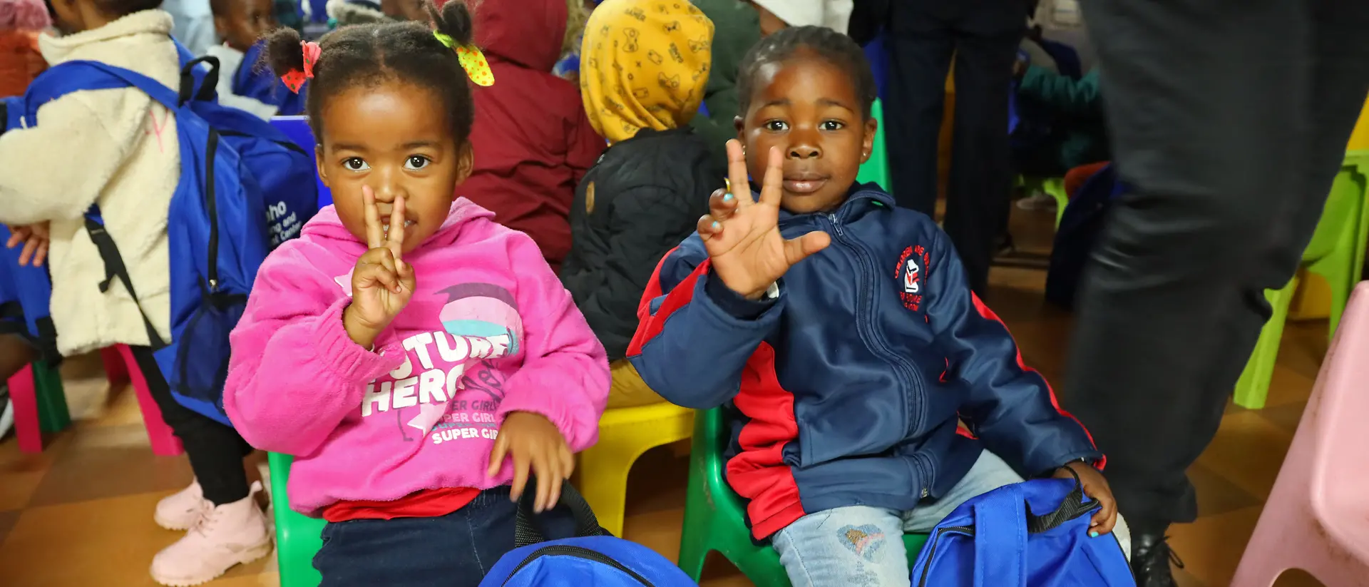 Two young children sit next to each other and hold up their hands.