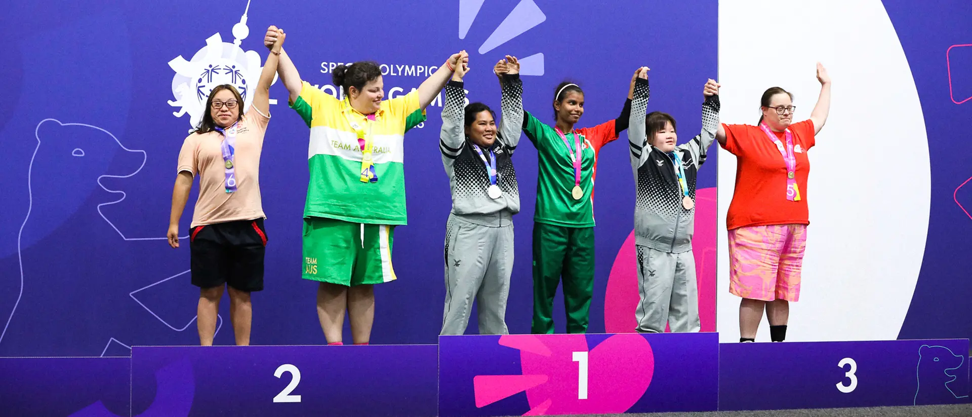 Six athletes stand on the podium at the Special Olympics and raise their arms in the air.