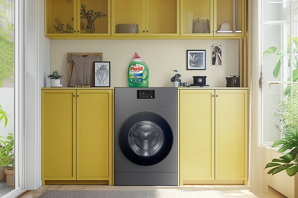 A Washing machine in the middle of a living room with Persil laundry detergent standing on top of it. 