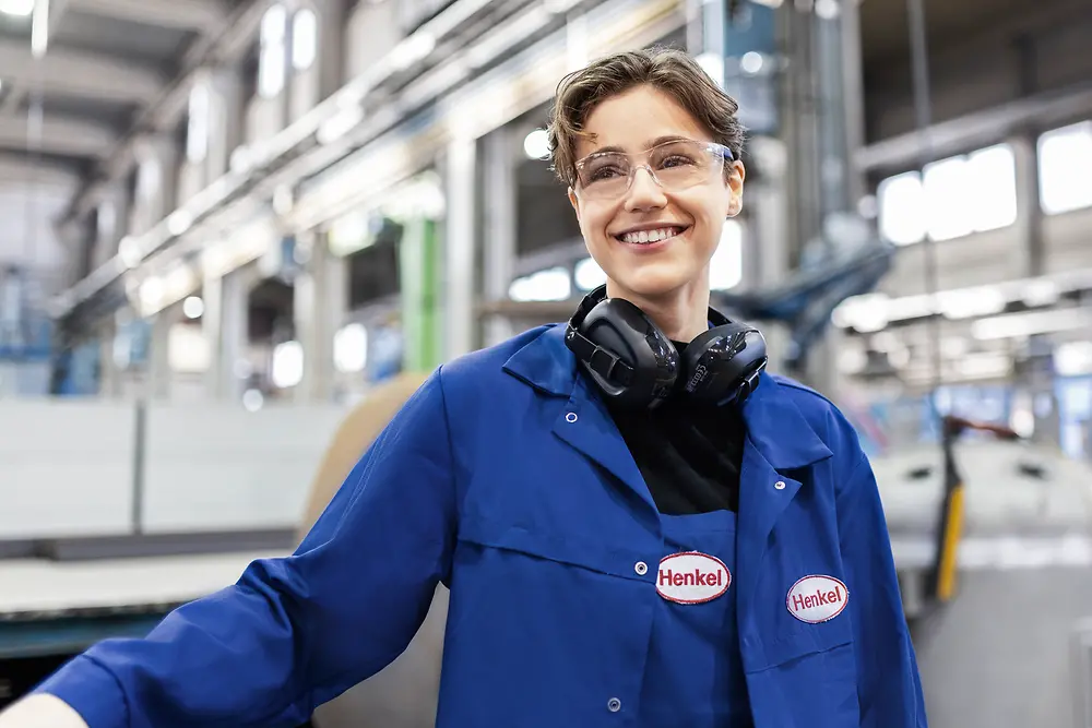 The photo shows a smiling young woman in a production hall wearing blue Henkel work clothes.