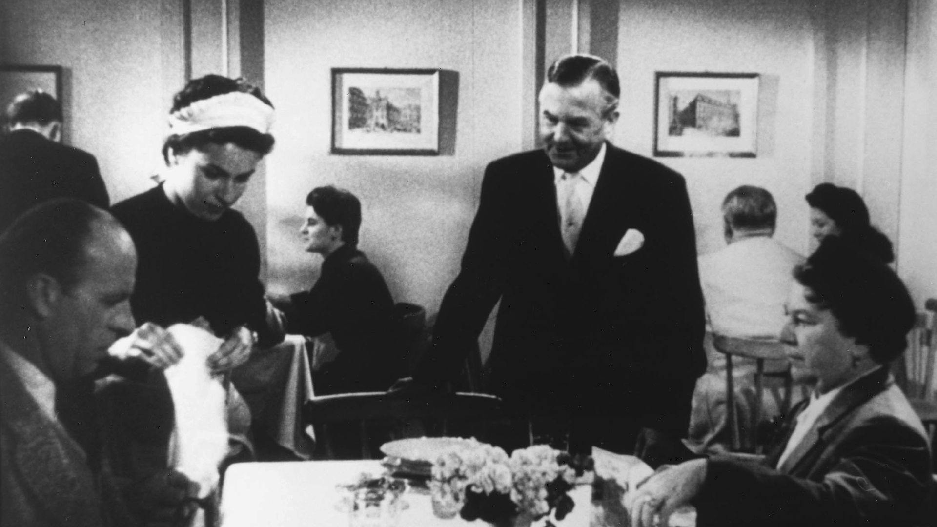 On the left a man and on the right a woman sitting at a restaurant table. Next to the man is a maid in a white bonnet. Next to the woman is a waiter.