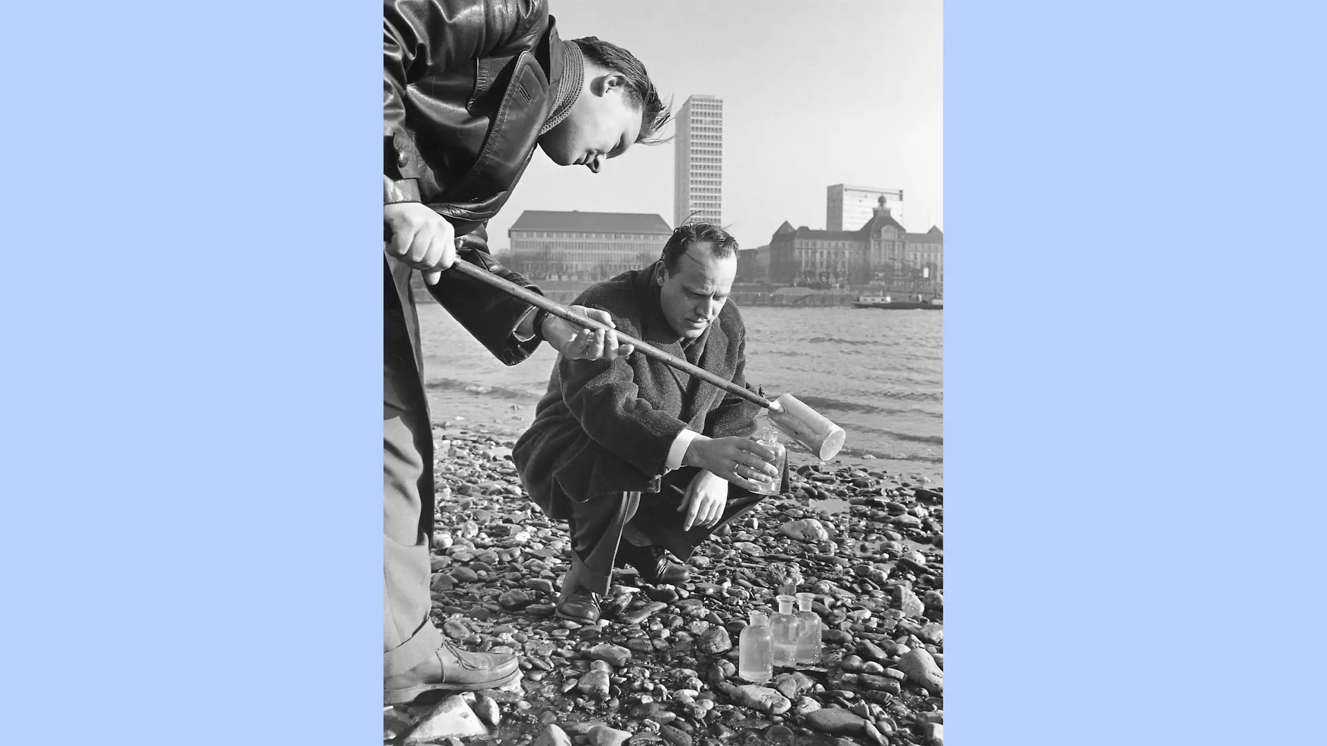 Black-and-white photograph of two men collecting a water sample from the river Rhine. One man is kneeling, holding a glass bottle, while the other bends down and fills the bottle using a wooden sampling stick.