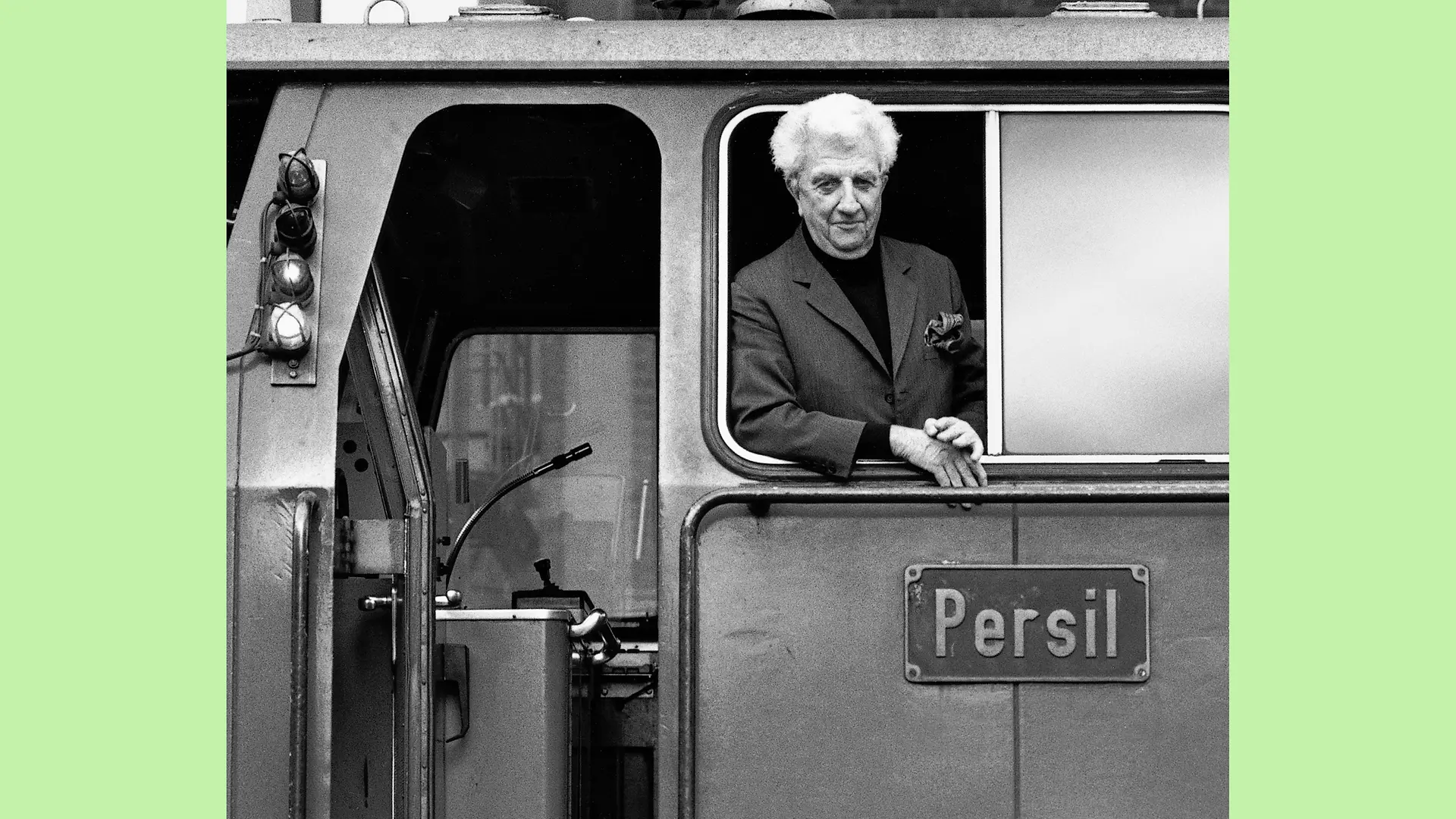 Dr. Konrad Henkel leans out of the side window of the driver’s cab of the locomotive Persil, smiling.