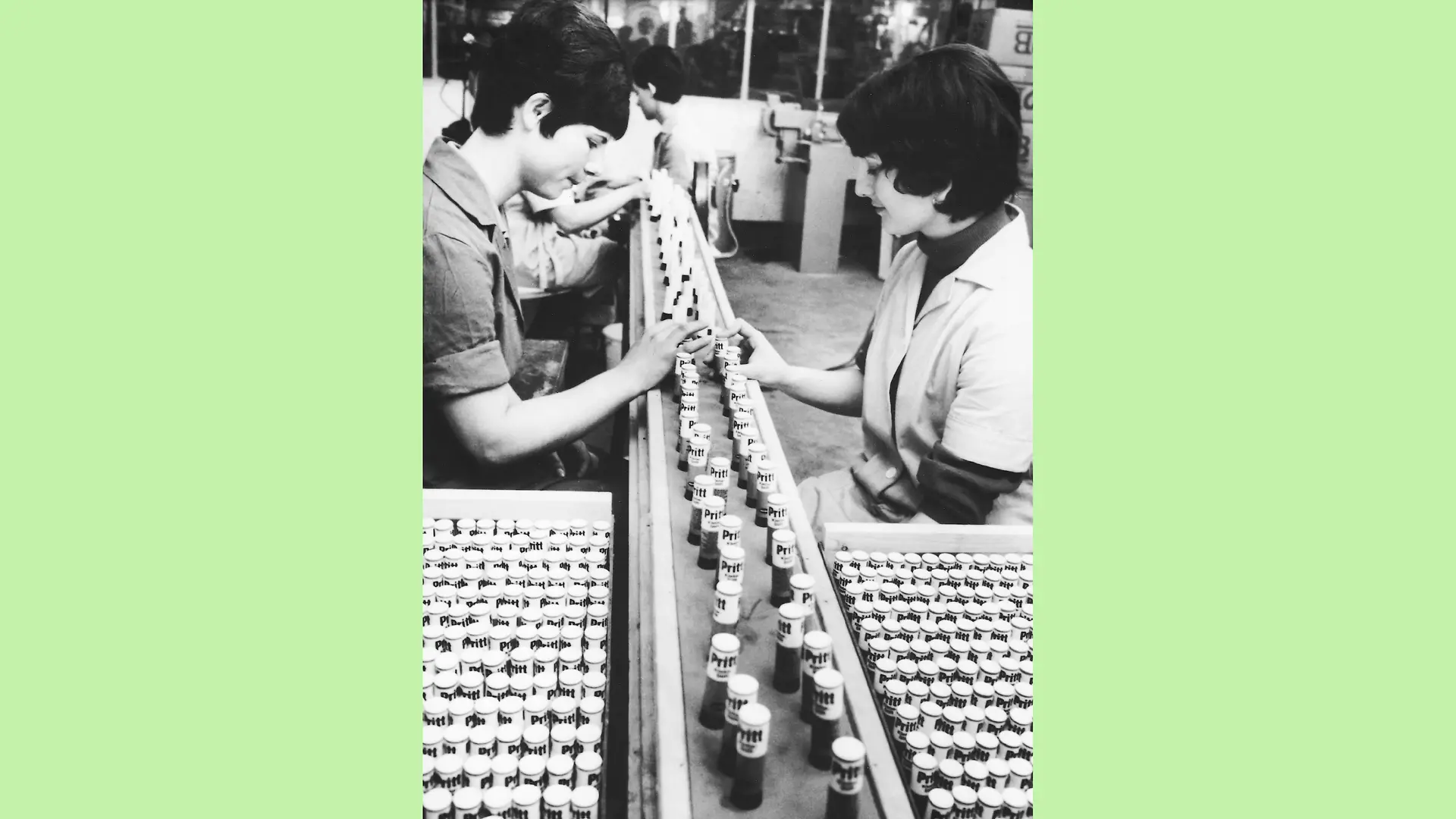 Black and white photo from the Pritt packaging plant. Two women are sitting opposite each other at a conveyor belt on which Pritt glue sticks are arranged in rows. Next to each woman is a box full of more Pritt glue sticks.