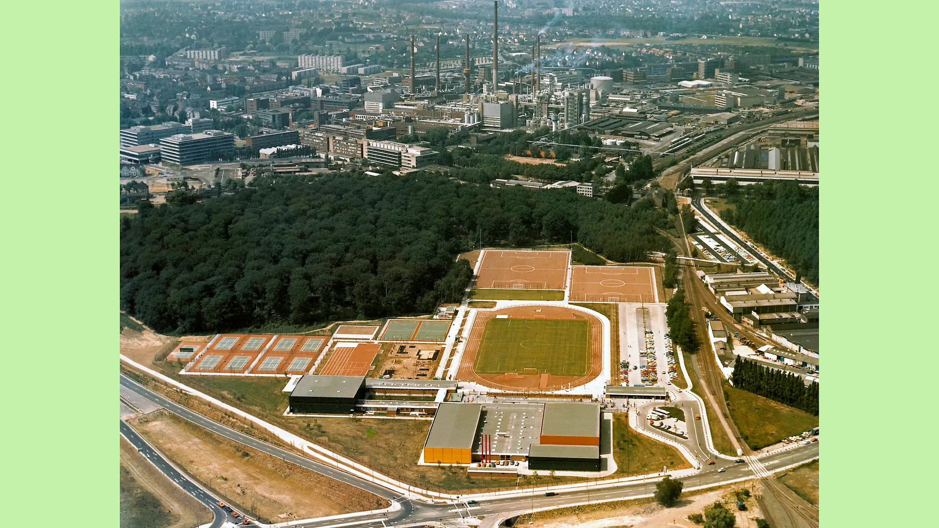 Aerial view of the Niederheid sports park. A grass football pitch with a running track forms the center of the sports park. Above it, slightly offset next to each other, are two further soccer pitches with a cinder surface. To the right of the grass pitch is the parking lot, half of which is filled with cars. Below the grass pitch is a hall that houses an indoor swimming pool. To the left of the hall is another building. To the left of the grass pitch are two handball courts, a playground and a total of nine tennis courts. In the background of the sports park, separated by a small wooded area, is the Henkel site.