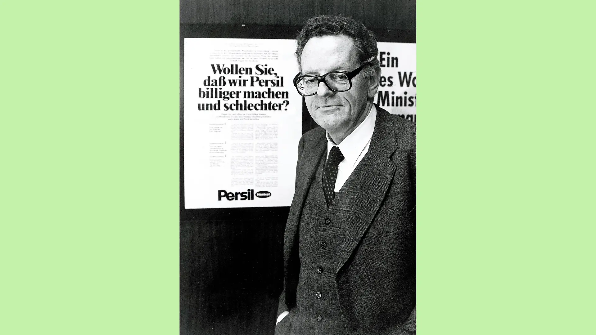 Black and white photo of Dr. Helmut Sihler in a dark three-piece suit and white shirt with a patterned tie. He is standing to the side, but facing the camera, next to a poster with the caption “Do you want us to make Persil cheaper and worse?”