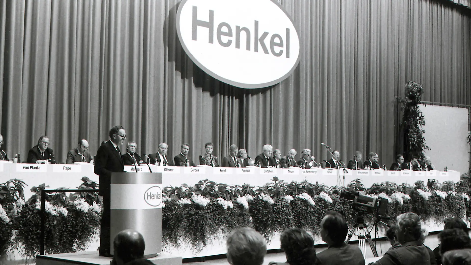 Dr. Dr. Helmut Sihler, the then Chairman of the Henkel Management Board, stands at the lectern and opens the first public Annual General Meeting in 1986. Behind him on stage sit the Management Board members and key company leaders of the time, positioned in front of their microphones.