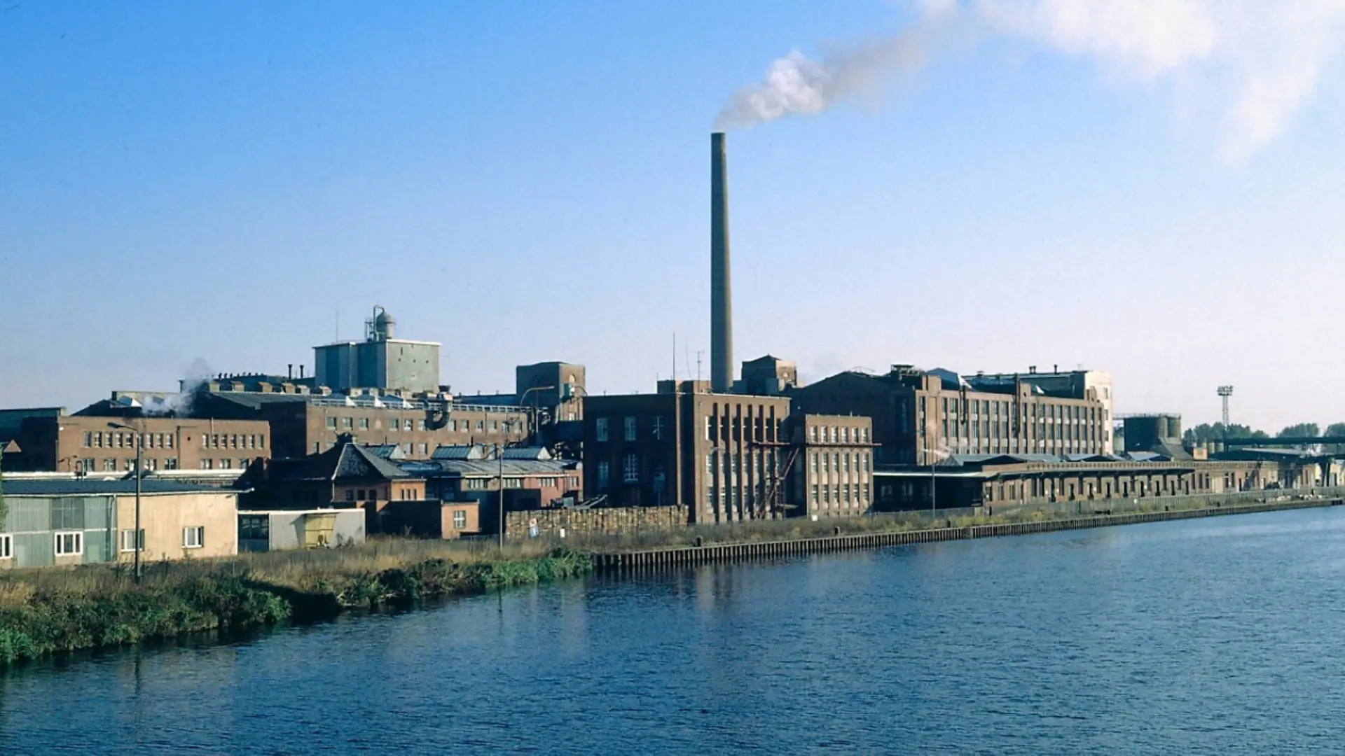 The Henkel plant in Genthin in the year it was bought back by Henkel (1990). The factory site borders a river. Many factory buildings can be seen. White smoke is rising from the chimney of one of the factory buildings.