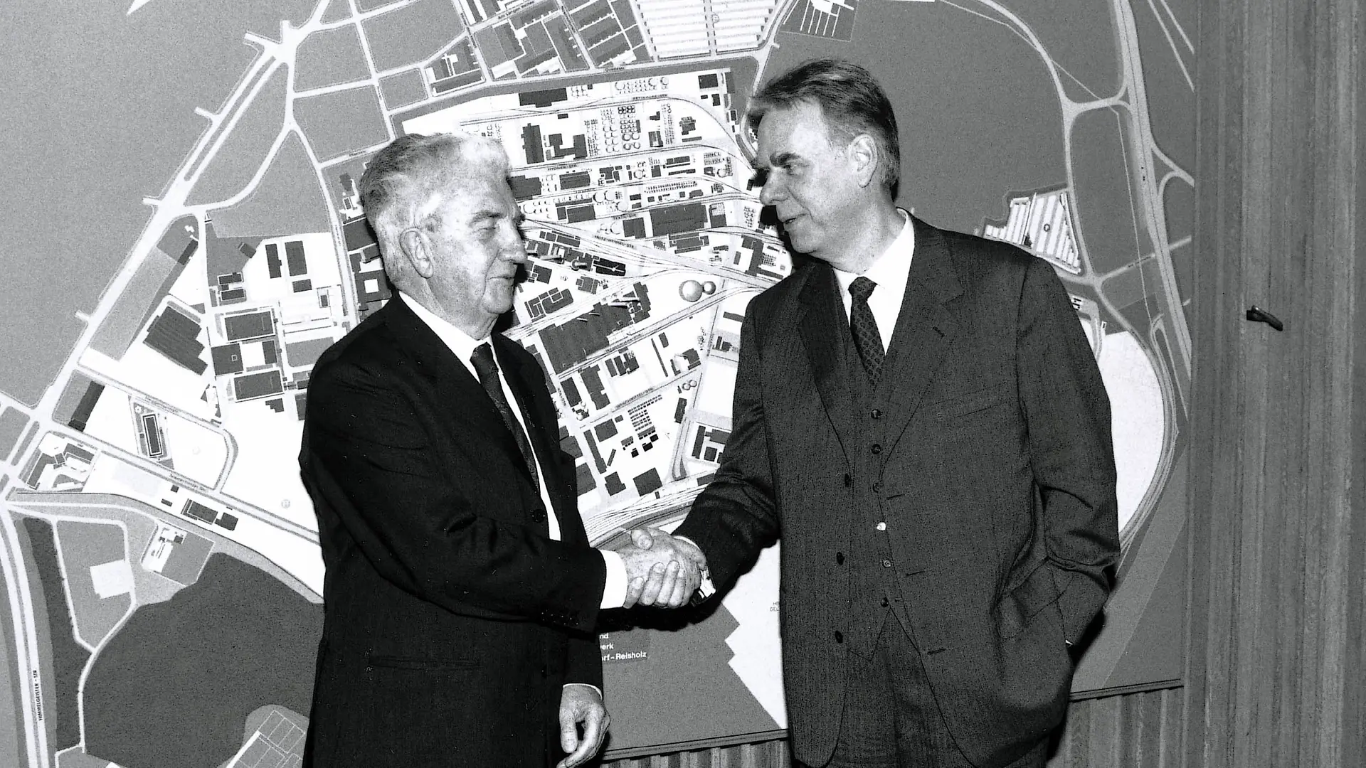 Dr. Konrad Henkel (left) and Dipl.-Ing. Albrecht Woeste face each other and shake hands. A large map of the plant in Düsseldorf-Holthausen hangs in the background.