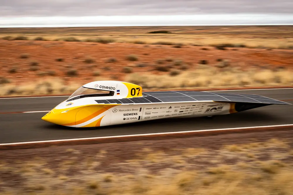 Yellow and white solar car “Aethon” with solar panels driving on a road through the red Australian Outback; sponsor logos visible, developed by a student team from Aachen and supported by Henkel adhesive technologies.