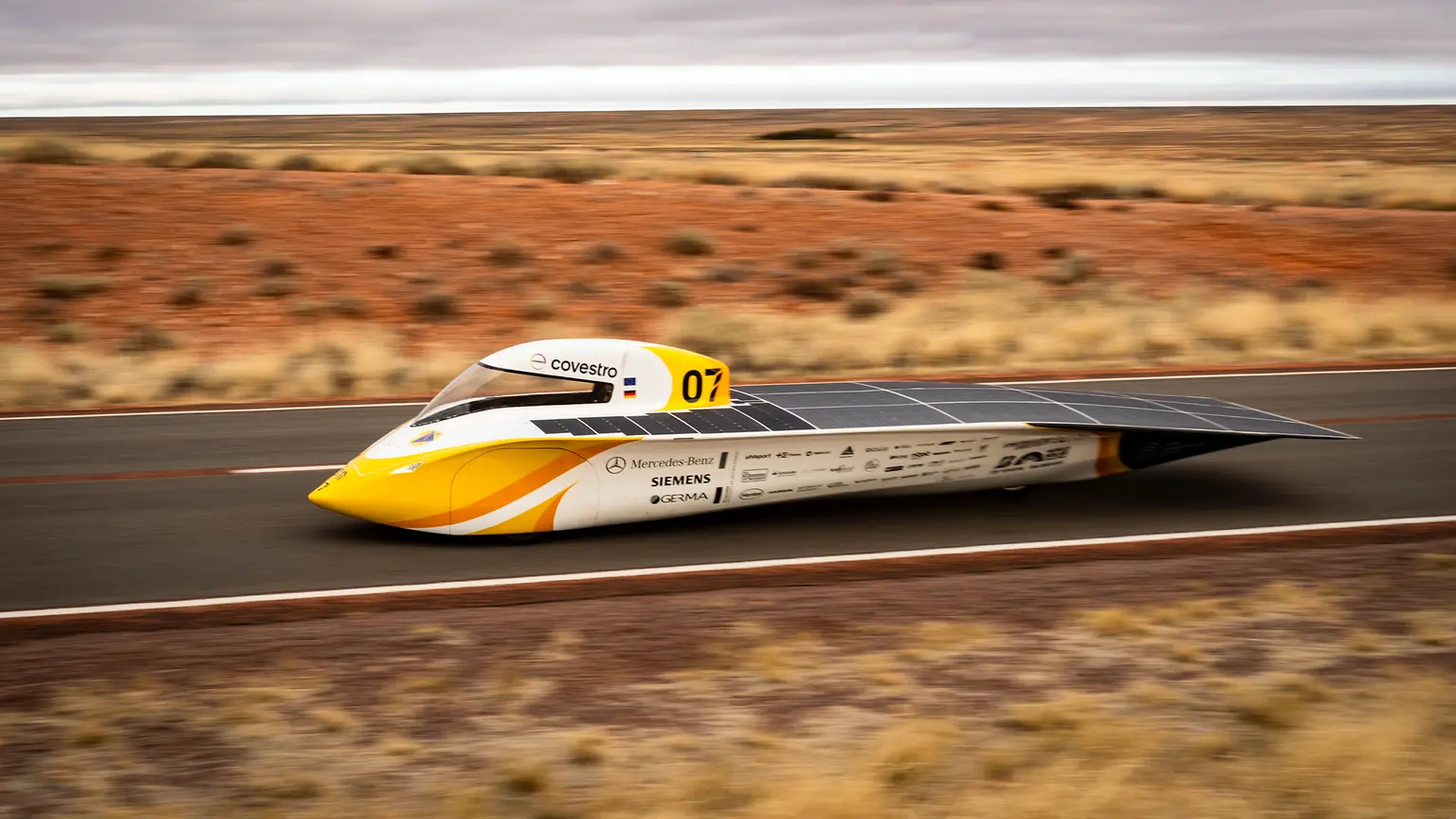 Yellow and white solar car “Aethon” with solar panels driving on a road through the red Australian Outback; sponsor logos visible, developed by a student team from Aachen and supported by Henkel adhesive technologies.