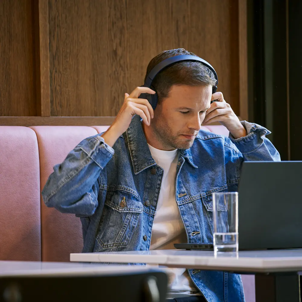 A man wearing a denim jacket and headphones is sitting at a table in a cafe, working on a laptop. There is a glass of water and a potted plant on the table.