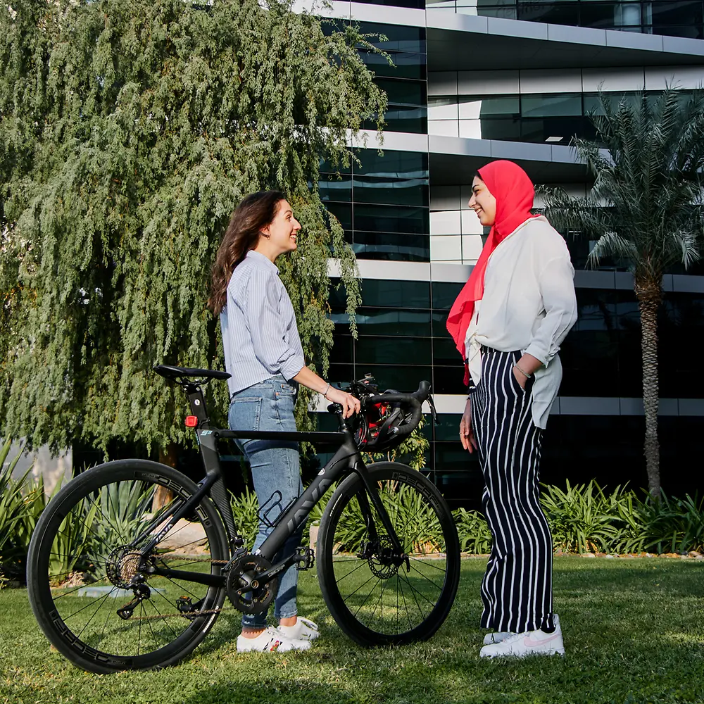 A photo of two women standing together in a green space in front of an office building with one of them holding her bike and helmet. 