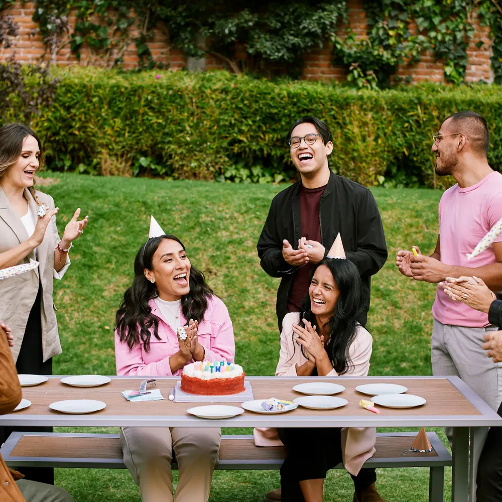 A group of eight colleagues wearing party hats standing in a garden around a desk celebrating the birthday of a woman and laughing together. 