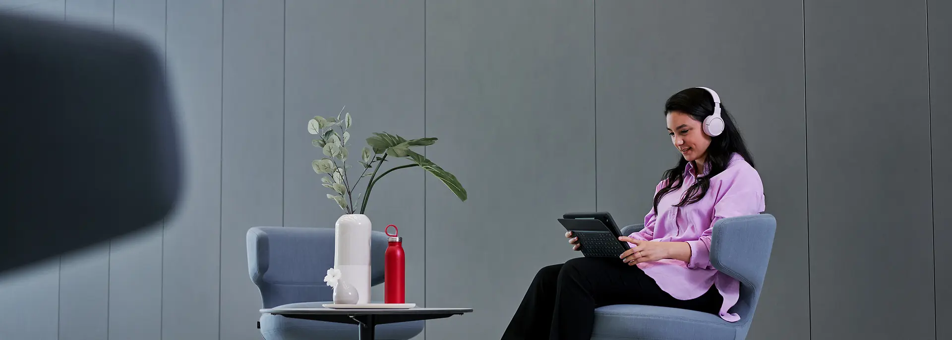 A photo of a woman sitting in a lounge chair focusing on her tablet device while wearing headphones.