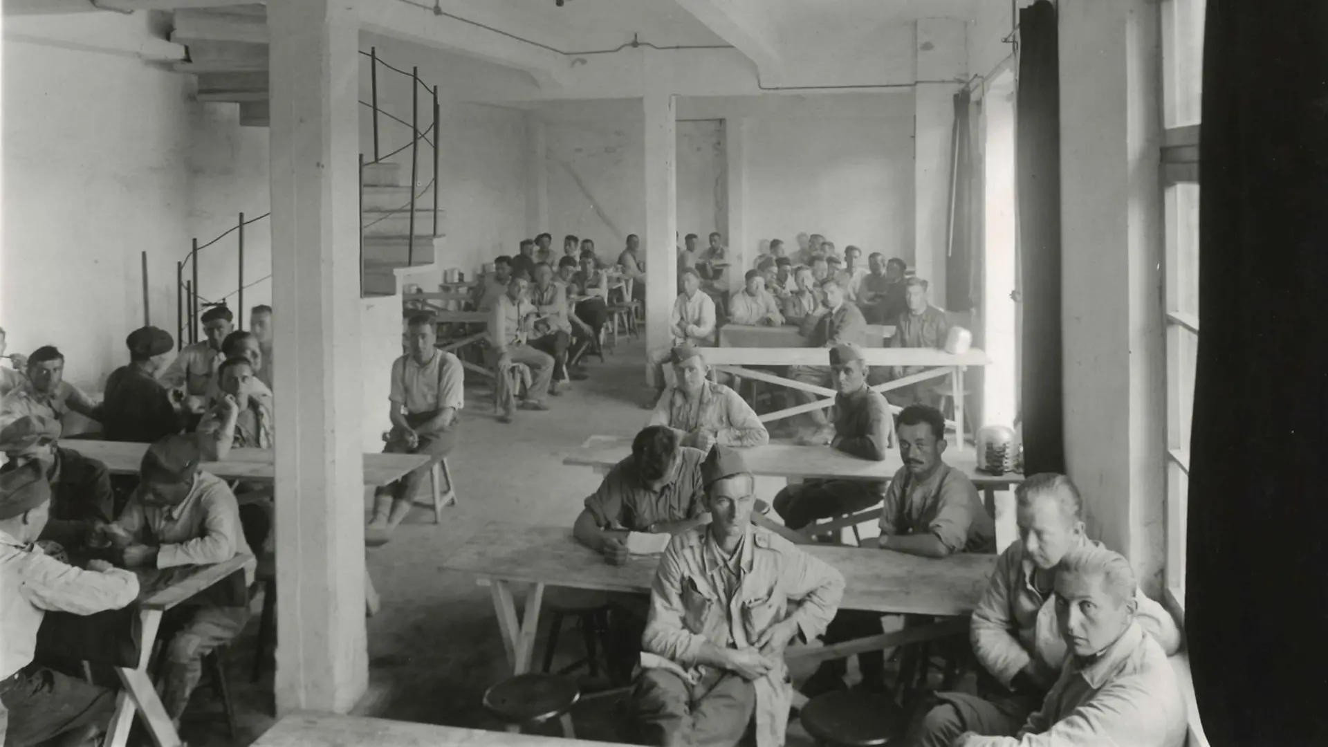 French prisoners of war in a common room with many tables and stools. Most of them look at the camera with a neutral expression. A spiral staircase leads into the room on the left-hand side.