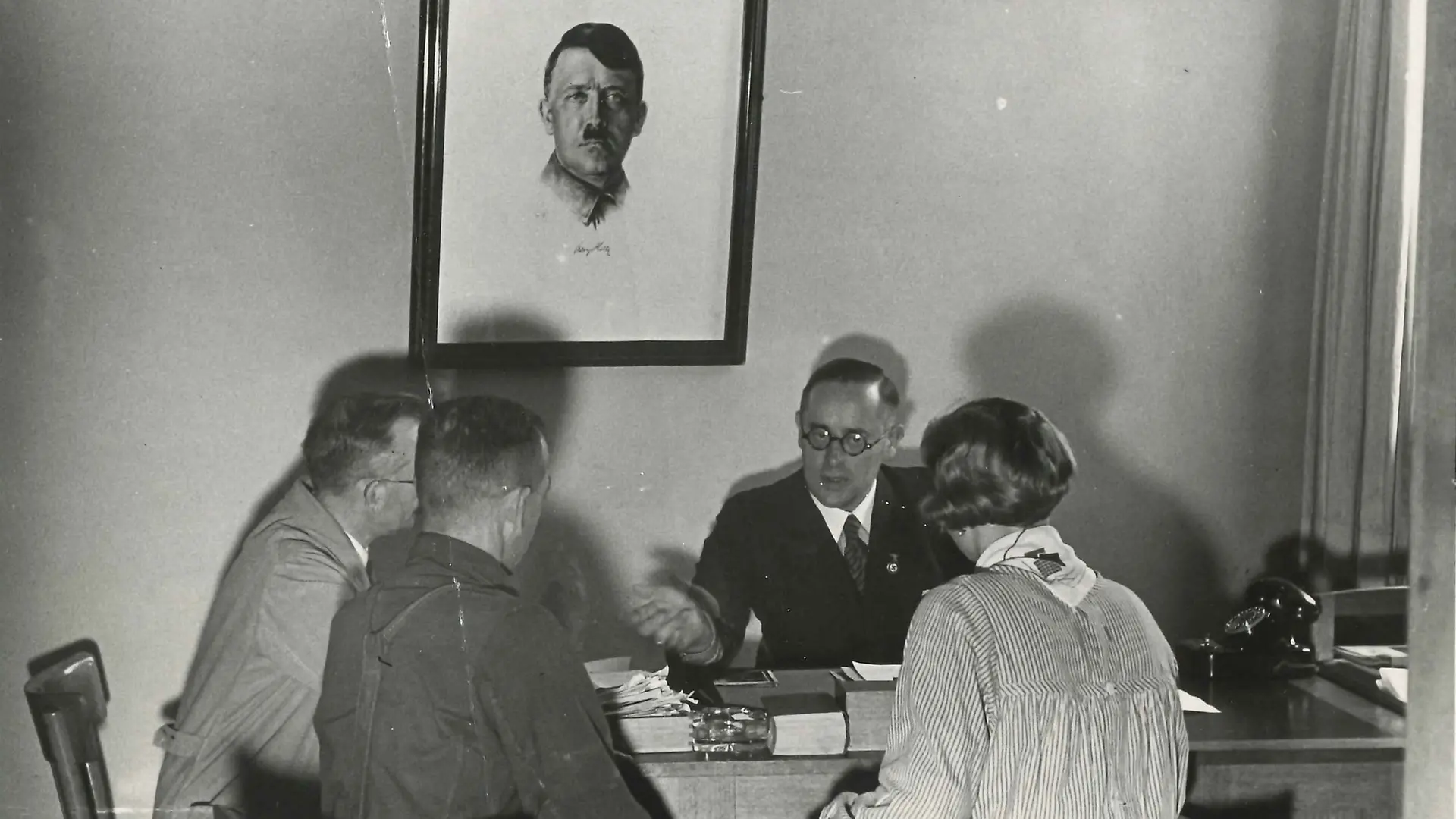 The four-member Henkel trust council, consisting of three men and one woman, gathered around a table. Two of the men and the woman are sitting with their backs to the camera. Above the table, on the wall, hangs a portrait of Adolf Hitler.