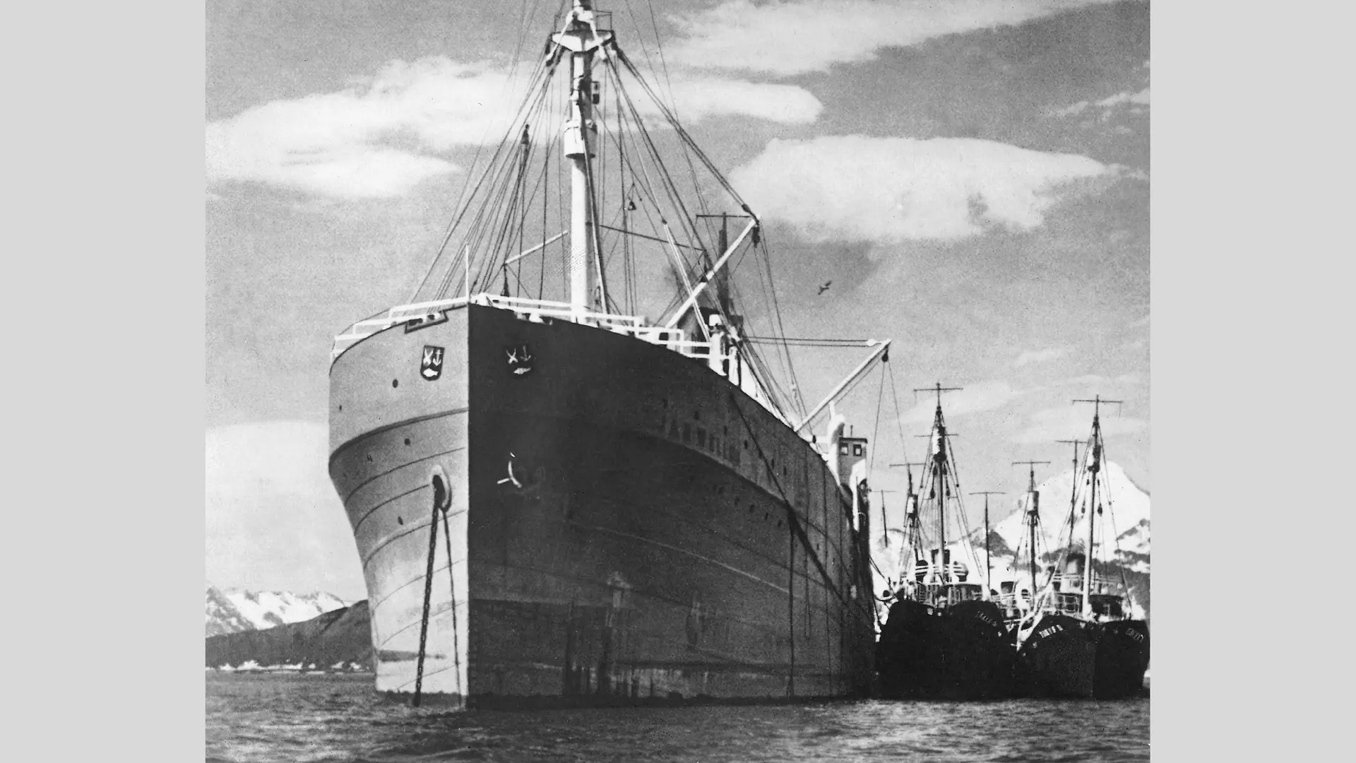 Black-and-white photograph of the whaling ship “Jan Wellem” moored in the harbor. The bow of the ship is visible in the center foreground. Several significantly smaller ships are docked to the right. Snow-capped mountains rise in the background under a partly cloudy sky.