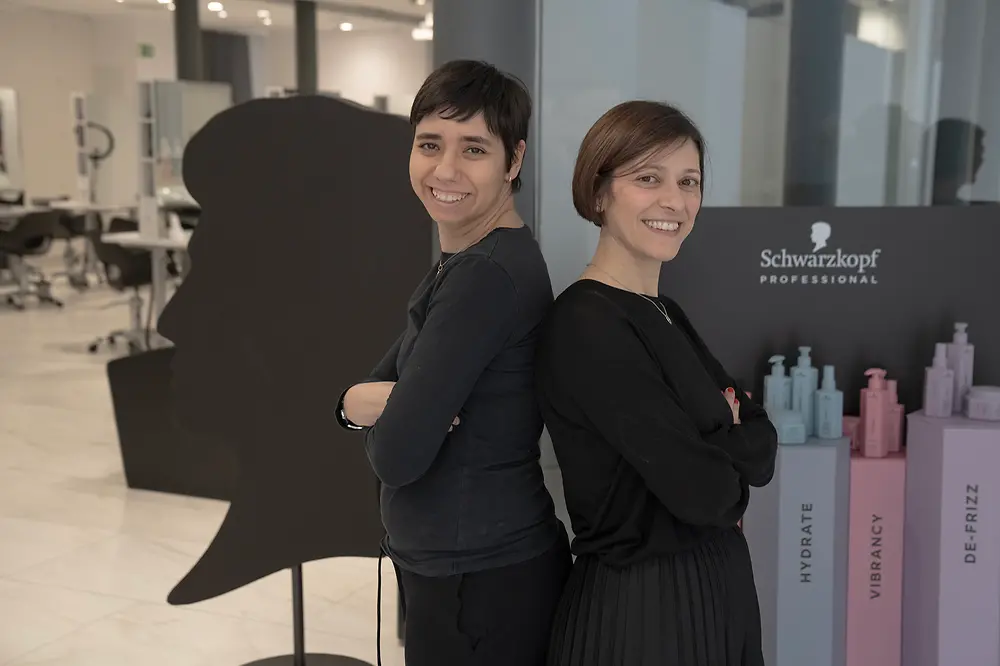 Noemí and her former trainer Meritxell stand back-to-back in a salon, smiling sideways toward the camera. 